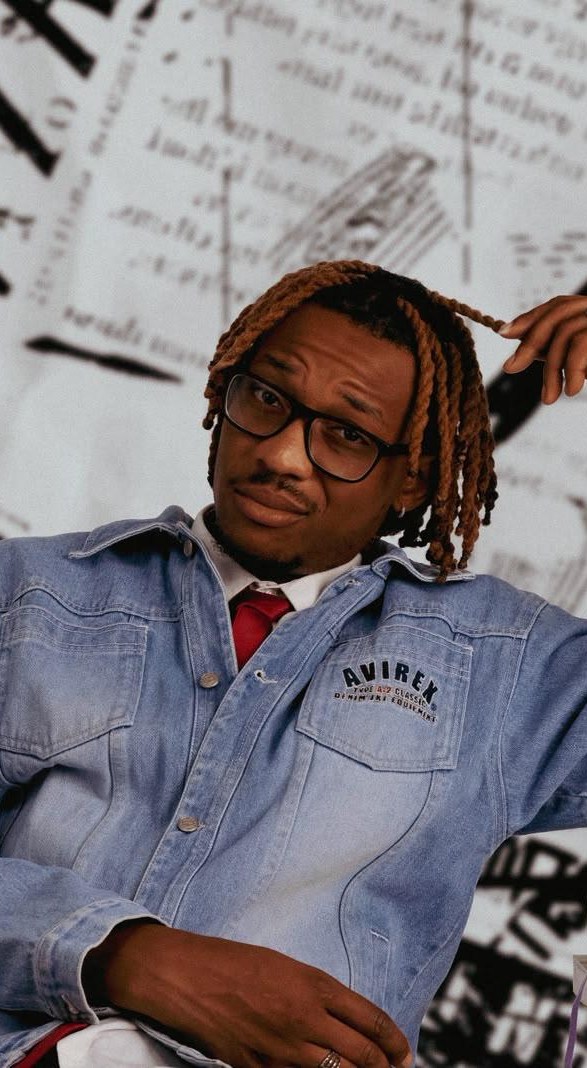 Mark Taiwo in an Avirex denim jacket and red tie, seated against a wall of newsprint, from the TROUBLE EP campaign shoot