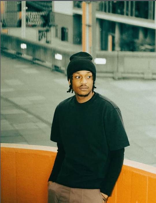 Mark Taiwo in a black beanie and black t-shirt, leaning against an orange wall in east London